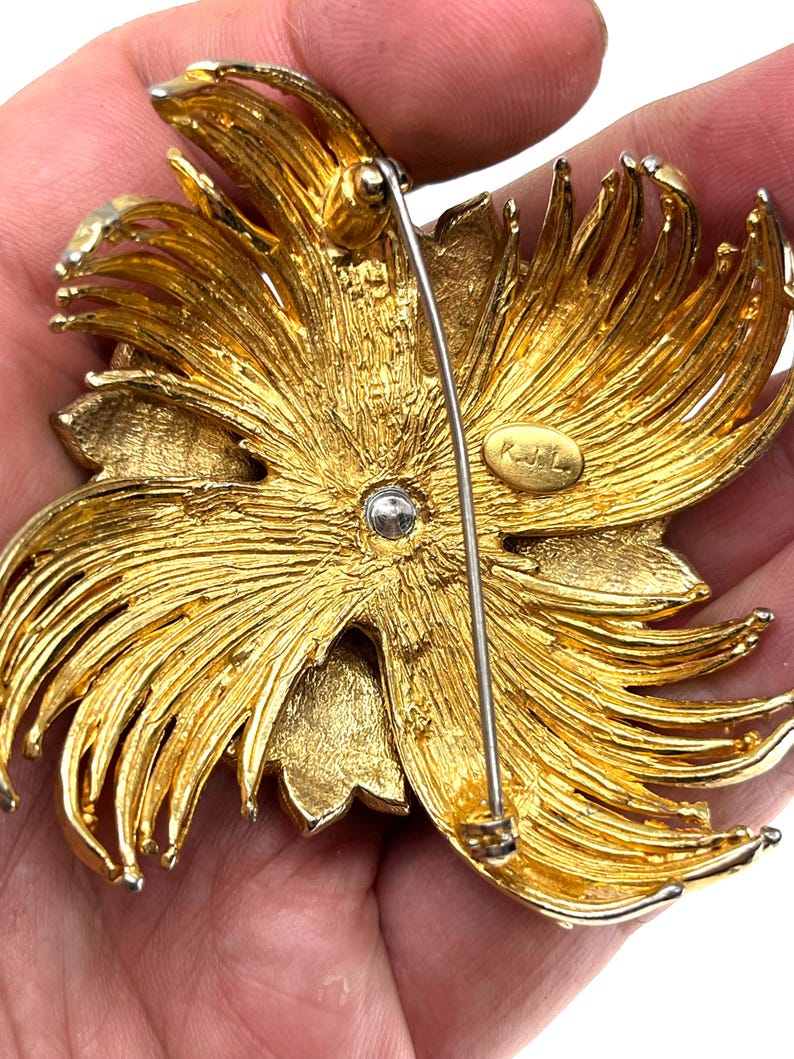 May include: A gold-toned metal brooch in a floral design. The brooch has a pin on the back for attachment. The design features layered petals and a textured finish. The brooch is likely vintage.