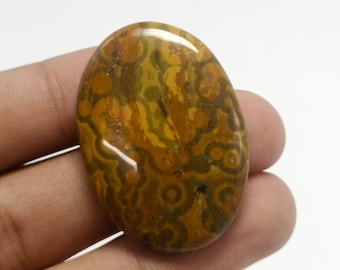 May include: Polished, oval gemstone with a pattern of swirling designs. The stone's colours include brown, yellow, and green. The gemstone is held in a hand against a white background.