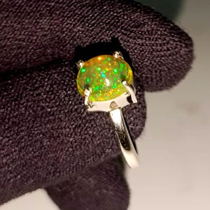 Puede incluir: Un anillo de plata con una gran piedra de ópalo ovalada. El ópalo muestra un juego de colores vibrante e iridiscente, con destellos verdes y amarillos. La banda del anillo es de plata pulida y sencilla.