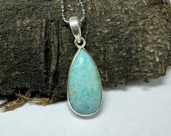 May include: A teardrop-shaped pendant with a light blue gemstone set in a silver bezel, suspended from a delicate silver chain. The gemstone has a smooth, polished surface. The pendant is displayed against a textured, dark brown wooden background.