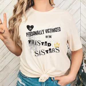May include: A white t-shirt with a black heart and the text "PERSONALLY VICTIMIZED BY THE TWISTED SISTERS" with a gold crown.