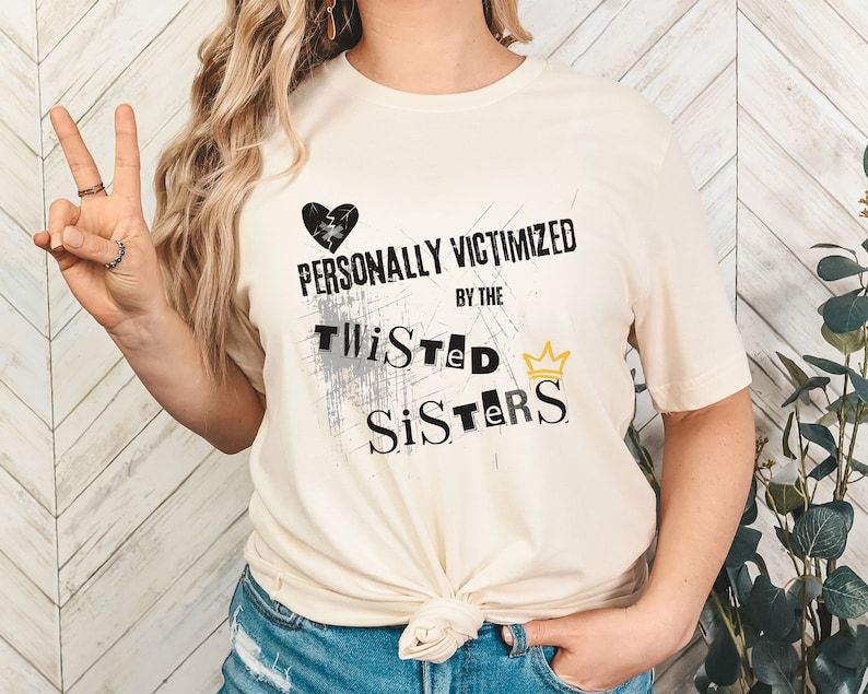 May include: A white t-shirt with a black heart and the text "PERSONALLY VICTIMIZED BY THE TWISTED SISTERS" with a gold crown.