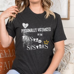 May include: A dark gray t-shirt with a white graphic that says "Personally Victimized by the Twisted Sisters" with a gold crown above the word "Sisters".