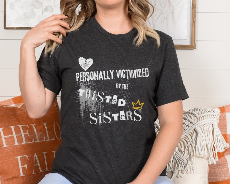 May include: A dark gray t-shirt with a white graphic that says "Personally Victimized by the Twisted Sisters" with a gold crown above the word "Sisters".