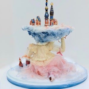 May include: A porcelain doll wearing a pink tulle skirt and a white blouse sits on a blue and white base. The doll is holding a miniature town on a cloud above her head. The town features several buildings with blue and red roofs.