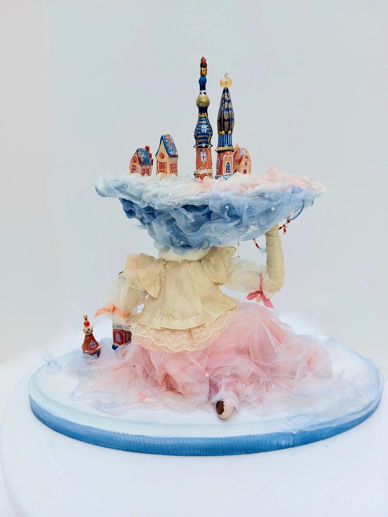 May include: A porcelain doll wearing a pink tulle skirt and a white blouse sits on a blue and white base. The doll is holding a miniature town on a cloud above her head. The town features several buildings with blue and red roofs.