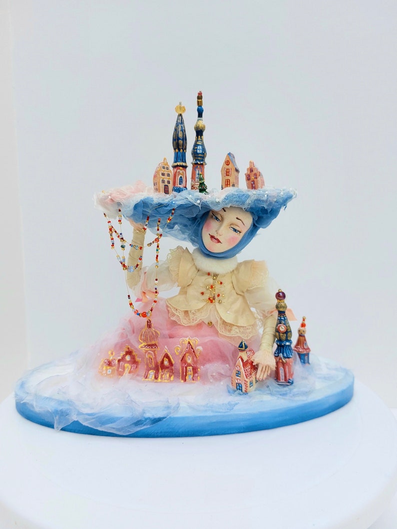 May include: A porcelain doll wearing a blue hat with a miniature city on top. The doll is sitting on a cloud-like base with more miniature houses and buildings. The doll has a pink dress and a beaded necklace.