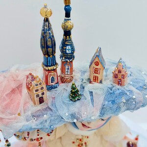 May include: A whimsical doll wearing a pink dress and a blue and white hat with miniature houses and a small Christmas tree on top. The doll has a sweet expression and is holding a small wand.