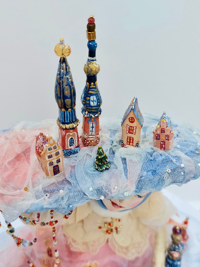 May include: A whimsical doll wearing a pink dress and a blue and white hat with miniature houses and a small Christmas tree on top. The doll has a sweet expression and is holding a small wand.