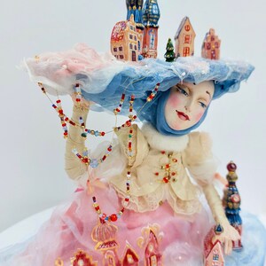 May include: A porcelain doll wearing a blue and white hat with a miniature town on top. The doll is wearing a white blouse, a pink skirt, and a blue scarf. The doll is holding a string of beads and a miniature house.