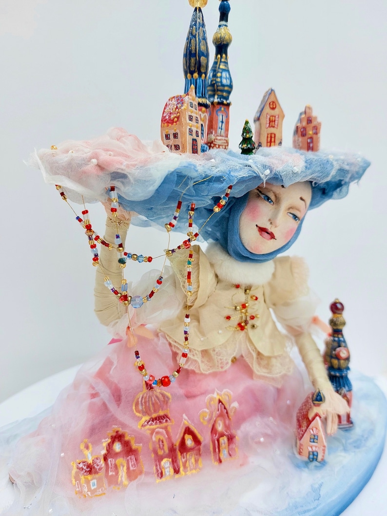 May include: A porcelain doll wearing a blue and white hat with a miniature town on top. The doll is wearing a white blouse, a pink skirt, and a blue scarf. The doll is holding a string of beads and a miniature house.