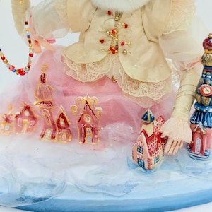 May include: A handmade doll wearing a cream-coloured jacket with gold accents and a pink skirt. The doll is holding a beaded string and standing on a snowy base with small painted houses.