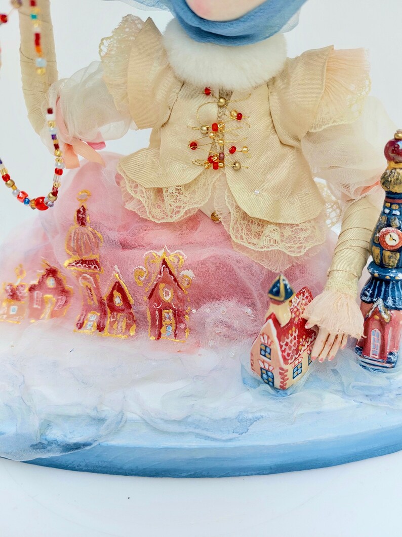 May include: A handmade doll wearing a cream-coloured jacket with gold accents and a pink skirt. The doll is holding a beaded string and standing on a snowy base with small painted houses.