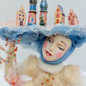 May include: A porcelain doll wearing a blue and white hat, adorned with miniature houses and a small Christmas tree. The doll is dressed in a white dress with pink accents.
