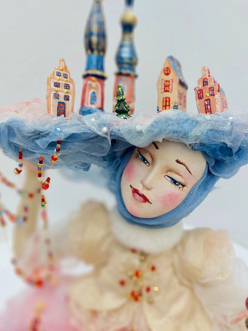 May include: A porcelain doll wearing a blue and white hat, adorned with miniature houses and a small Christmas tree. The doll is dressed in a white dress with pink accents.