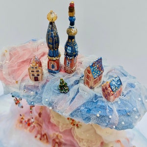 May include: A miniature winter village scene featuring blue and white houses, a small Christmas tree, and two tall blue and gold spires. The village is set on a fluffy white cloud with pink and blue accents.