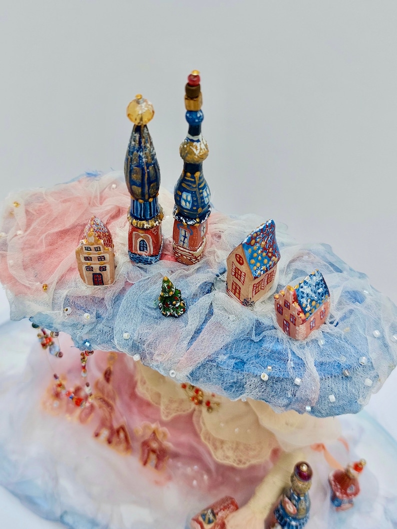 May include: A miniature winter village scene featuring blue and white houses, a small Christmas tree, and two tall blue and gold spires. The village is set on a fluffy white cloud with pink and blue accents.