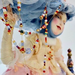May include: A porcelain doll wearing a blue and white hat, a pink dress and a beaded necklace with red, blue and gold beads.