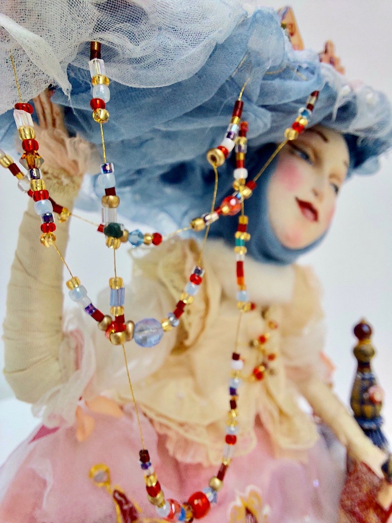 May include: A porcelain doll wearing a blue and white hat, a pink dress and a beaded necklace with red, blue and gold beads.