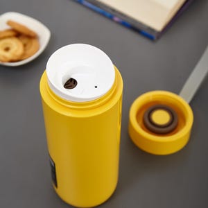 May include: A yellow thermos with a white lid and a small hole in the lid. The thermos is sitting on a gray surface.