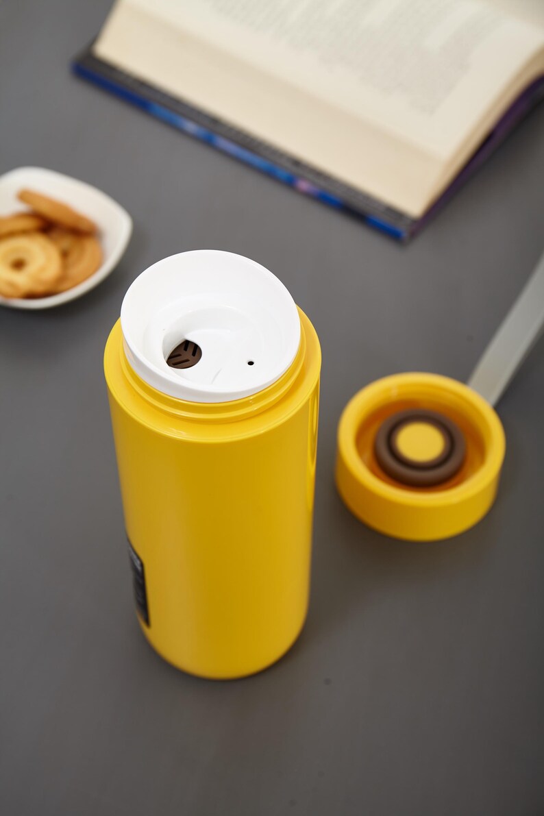 May include: A yellow thermos with a white lid and a small hole in the lid. The thermos is sitting on a gray surface.