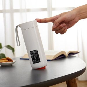 May include: White travel mug with a black handle and a black label with instructions on how to use the mug. The mug is sitting on a white table.