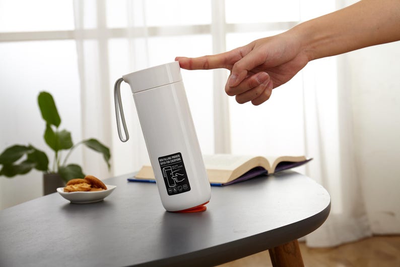 May include: White travel mug with a black handle and a black label with instructions on how to use the mug. The mug is sitting on a white table.