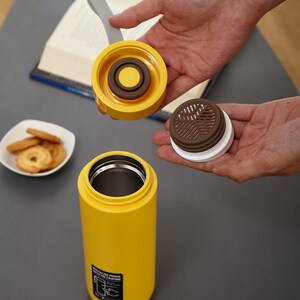 May include: A yellow stainless steel water bottle with a brown and white lid. The lid has a removable filter. The bottle has a black label that says "Double Power Keep It Fresh For Everyone".