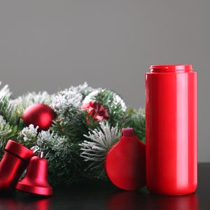 May include: A red insulated travel mug with a lid, sitting on a black surface next to a green Christmas wreath with red ornaments and bells.
