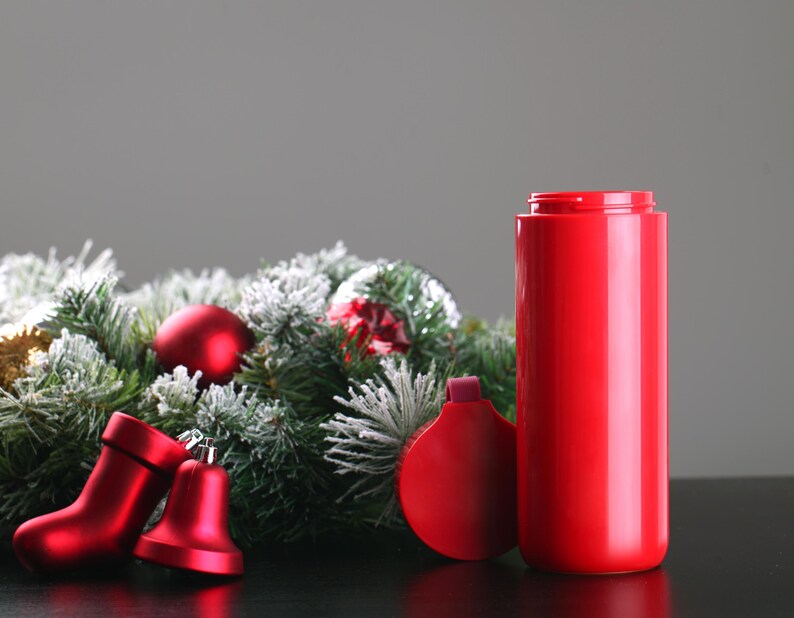 May include: A red insulated travel mug with a lid, sitting on a black surface next to a green Christmas wreath with red ornaments and bells.