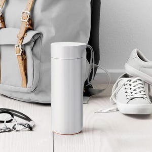 May include: A white stainless steel water bottle with a black carrying strap. The bottle is sitting on a light wood surface next to a gray backpack and white sneakers.