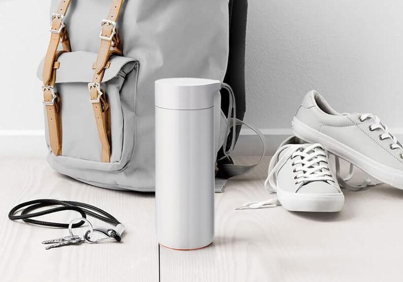 May include: A white stainless steel water bottle with a black carrying strap. The bottle is sitting on a light wood surface next to a gray backpack and white sneakers.