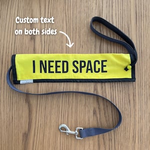 May include: Yellow dog lead with the text "I NEED SPACE" in black. The lead has a black border and a grey handle and lead. A silver-coloured clasp is attached to the lead.