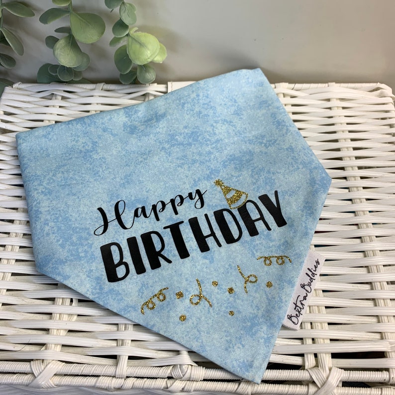 May include: A light blue bandana with the text "Happy Birthday" in black lettering and a gold glitter party hat. The bandana has gold glitter confetti and a white tag with the text "Bostra Bulldogs" in black lettering.
