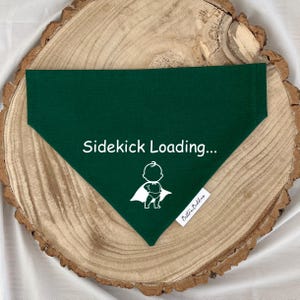 K&ouml;nnte beinhalten: Ein dunkelgr&uuml;nes, dreieckiges Haustierhalstuch mit dem wei&szlig;en Schriftzug "Sidekick Loading..." &uuml;ber einer wei&szlig;en Grafik eines Babys mit Superheldenumhang. An der unteren rechten Kante befindet sich ein kleines wei&szlig;es Stoffetikett mit "Bestie Buddies".
