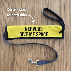May include: A yellow dog leash sleeve with black trim and the black text NERVOUS GIVE ME SPACE printed on the front. The sleeve features a loop handle and a metal swivel snap hook at the end. The text Custom text on both sides is visible.