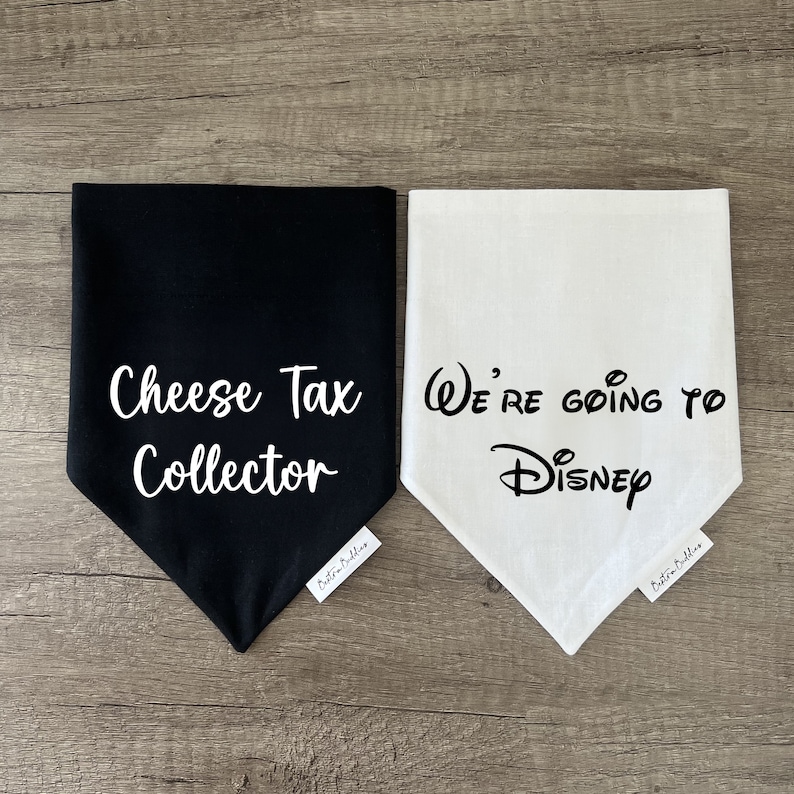 K&ouml;nnte beinhalten: Zwei dreieckige Haustierhalst&uuml;cher, eines in Schwarz mit wei&szlig;em Text Cheese Tax Collector und eines in Wei&szlig; mit schwarzem Text We're going to Disney. Jedes Halstuch hat ein kleines wei&szlig;es Stoffetikett mit dem Markennamen Bestie Buddies.