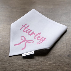 May include: White bandana with the name "Harley" printed in pink cursive lettering. A pink bow is centered above the name. The bandana has a small white tag with the text "Boutique Buddies" printed in black.