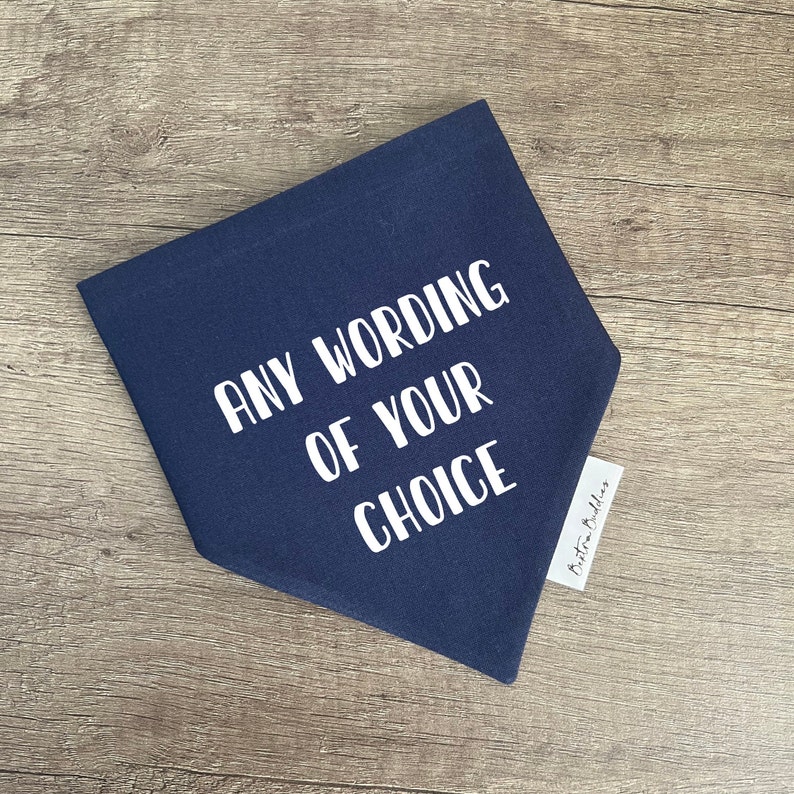 K&ouml;nnte beinhalten: Ein dreieckiges Bandana aus marineblauem Stoff mit dem wei&szlig;en Text ANY WORDING OF YOUR CHOICE in einer serifenlosen Schriftart. Ein kleines wei&szlig;es Stoffetikett mit dem schwarzen Text Bexton Buddies ist an der rechten Seite des Bandanas befestigt.