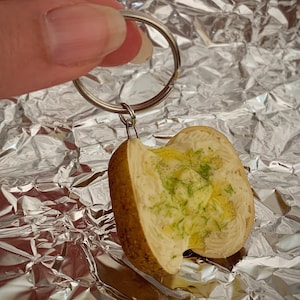 May include: A miniature keyring shaped like a baked potato cut in half. The potato features a textured brown skin and a creamy white interior topped with a pat of yellow butter, green herbs, and black pepper, attached to a silver metal ring.