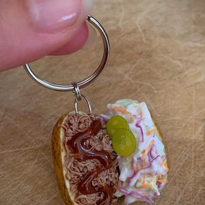 May include: A miniature food-themed keyring featuring a halved baked potato. One side is topped with shredded brown meat drizzled with dark brown sauce, while the other side features creamy white coleslaw with orange and purple shreds and two green pickle slices.