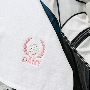 May include: White golf towel with a pink embroidered golf ball and wreath with the word "DANY" underneath. The towel is hanging on a white and black golf bag.