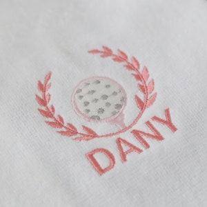 May include: White golf towel with pink embroidery of a golf ball and leaves, with the name "DANY" embroidered below.