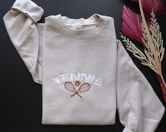 Puede incluir: Una sudadera beige con la palabra "TENNIS" bordada en blanco con dos raquetas de tenis marrones cruzadas debajo.