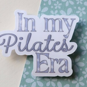 May include: White sticker with the text "In my Pilates Era" in a light blue color with a snowflake pattern.