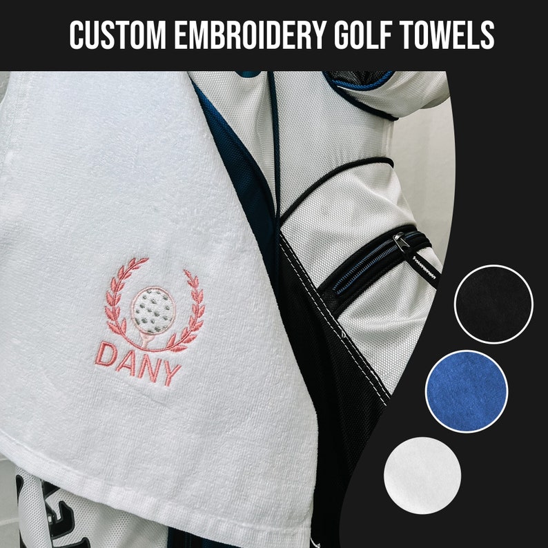 May include: White golf towel with pink embroidery of a golf ball inside a wreath with the word "DANY" below. The towel is hanging on a golf bag. Three color swatches are shown: black, blue, and white.