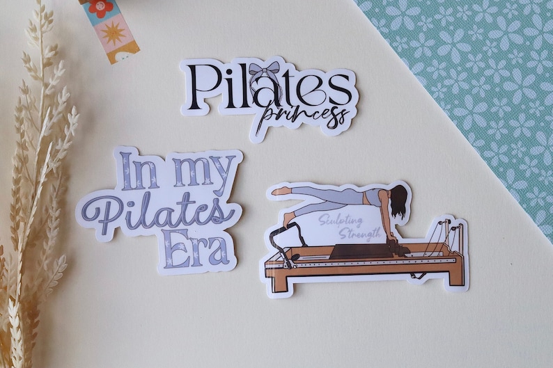 May include: Three Pilates-themed stickers. One sticker features a woman doing Pilates on a reformer with the text "Sculpting Strength". Another sticker says "In my Pilates Era" in a light blue font. The third sticker says "Pilates princess" in black font with a gray bow.
