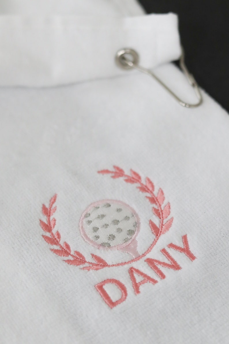 May include: White golf towel with pink embroidery of a golf ball and leaves, with the name "DANY" embroidered below.