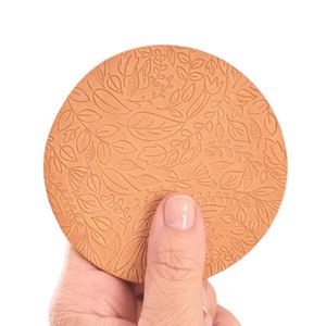 May include: A round, brown leather coaster with a floral pattern. The coaster is being held by a hand.