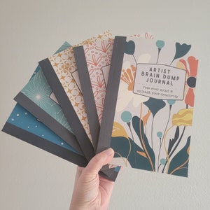 May include: A stack of five journals with different floral and star patterns. The top journal has a floral design and the text "Artist Brain Dump Journal" with the tagline "free your mind & unleash your creativity".